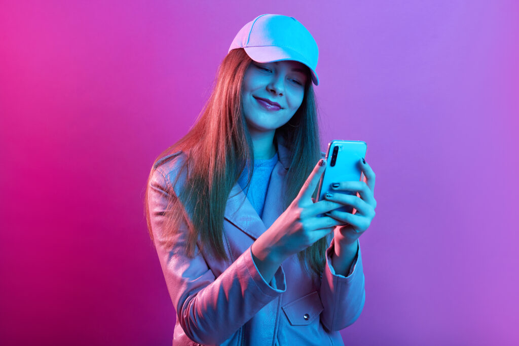 Portrait of young beautiful fashionable model wearing leather baker and baseball cap, holding smart phone in hands, texting with friends or checking social networks.
